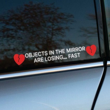 Plotrovaná nálepka OBJECTS IN THE MIRROR ARE LOSING... FAST s červenými zlomenými srdcami pre výrazný kontrast.