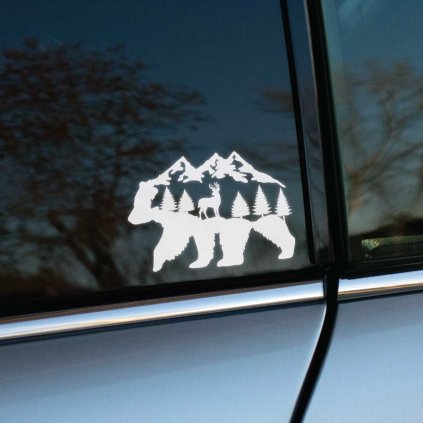 Plotrovaná nálepka v siluete kráčajúceho medveďa, v ktorej je vyobrazený jeleň medzi stromami pod štítmi vysokých hôr.