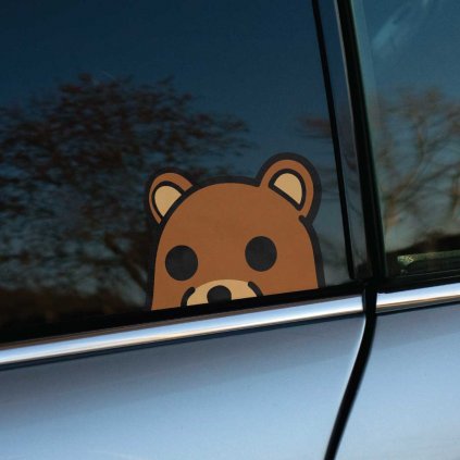 Tlačená nálepka na auto s hlavou kresleného medvedíka, ktorý vykúka zospodu okna, jednoduchý a rozpoznateľný dizajn.
