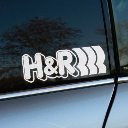 Nálepka s logom „H&R“ nalepená na zadnom bočnom okne auta. Výrazný a športový dizajn s charakteristickými tromi vlnami symbolizuje kvalitu a výkon zameraný na úpravy podvozkov. Lesklé sklo s odrazmi okolitej prírody zvýrazňuje precíznosť aplikácie nálepky a jej estetický vzhľad. Ideálna voľba pre automobilových nadšencov a milovníkov detailov, ktoré podčiarkujú jedinečnosť vozidla.