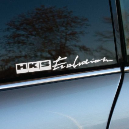 Biela nálepka „HKS Evolution“ kombinuje ikonické štvorcové logo „HKS“ s elegantným rukopisným nápisom „Evolution“, nalepená na tmavom bočnom okne auta. Tento dizajn spája precíznosť a dynamiku, čím odráža záväzok značky HKS k inováciám v oblasti vysokovýkonného tuningu. Kontrast medzi ostrými geometrickými tvarmi loga a plynulými líniami textu vytvára vizuálne zaujímavý prvok, ktorý vyniká na tmavom pozadí. Strieborná karoséria auta dokonale dopĺňa sofistikovaný a športový vzhľad. Kľúčové slová: nálepka HKS Evolution, vysokovýkonné diely, tuningové doplnky, športové auto, nálepky na auto, personalizácia vozidla.
