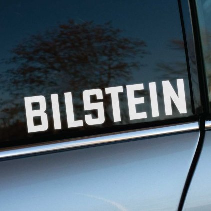 Biela nálepka „BILSTEIN“ nalepená na bočnom okne tmavosivého automobilu. Elegantné prevedenie písma vyniká na čistej sklenej ploche a odráža okolitú krajinu. Ideálna nálepka pre fanúšikov automobilových značiek a tuningových doplnkov. Skvelý spôsob, ako personalizovať auto a zdôrazniť záľubu v kvalitných tlmičoch Bilstein. Perfektné pre milovníkov štýlových a funkčných nálepiek na vozidlá.