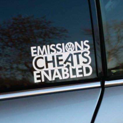 Plotrovaná nálepka s textom „EMISSIONS CHEATS ENABLED“ a logom Volkswagen na bočnom okne auta.