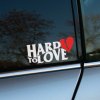 Plotrovaná nálepka na auto s výrazným nápisom „HARD TO LOVE“ a červeným rozpoleným srdcom ako symbolom emócií.