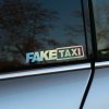Plotrovaná nálepka s holografickým odleskom, ktorú tvorí nápis v obdĺžnikovom ráme s textom v znení FAKE TAXI.