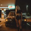 Upravené BMW E36 pri čerpacej stanici v noci, pred autom stojí žena v čiernom tope a lesklých šortkách.
