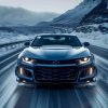 Predný pohľad na elegantné a výkonné Chevrolet Camaro, ktoré sa rýchlo pohybuje po zasneženej ceste v horskom prostredí. Auto vyžaruje silu a dynamiku, pričom jeho ostré línie a agresívny dizajn dokonale ladia s nálepkou lebky na čelnom skle. Zasnežená krajina a kľukaté cesty v pozadí vytvárajú dramatický kontrast s lesklým povrchom vozidla, zdôrazňujúc jeho schopnosti a štýl. Tento obraz je ideálny pre milovníkov športových áut, ktorí oceňujú nielen výkon, ale aj estetiku a jedinečný dizajn vozidiel značky Chevrolet.