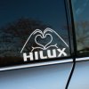 Na obrázku je nálepka na bočnom okne auta Toyota Hilux. Dve ruky formujúce srdce sú umiestnené nad nápisom "HILUX", čo dodáva tejto nálepke osobitý a výrazný vzhľad. Tento kreatívny dizajn je ideálny pre všetkých majiteľov a fanúšikov modelu Toyota Hilux, ktorí chcú svojmu vozidlu pridať unikátny a osobný dotyk. Nálepka nielen zlepšuje vizuálny vzhľad auta, ale tiež vyjadruje vašu vášeň pre tento robustný a spoľahlivý model. Je to perfektný spôsob, ako prejaviť svoju lásku k Toyote Hilux a spraviť svoje auto ešte výnimočnejším na cestách aj v teréne.