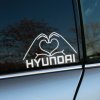 Na obrázku je nálepka na bočnom okne auta Hyundai. Dve ruky formujúce srdce sú umiestnené nad nápisom "HYUNDAI", čo dodáva tejto nálepke osobitý a vkusný vzhľad. Tento kreatívny dizajn je ideálny pre všetkých majiteľov a fanúšikov značky Hyundai, ktorí chcú svojmu vozidlu pridať unikátny a osobný dotyk. Nálepka nielen zlepšuje estetiku vášho auta, ale tiež vyjadruje vašu vášeň pre túto spoľahlivú a populárnu automobilovú značku. Perfektný spôsob, ako prejaviť lásku k Hyundai a spraviť svoje auto výnimočným na cestách.
