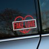 Plotrovaná nálepka na auto so zlomeným srdcom a výrazným nápisom „FAKE LOVE“ v šikmom obdĺžnikovom rámčeku.