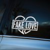 Plotrovaná nálepka na auto so zlomeným srdcom v pozadí a nápisom „FAKE LOVE“ v šikmo osadenom rámčeku.