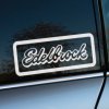 Biela samolepka s logom "Edelbrock" na okne auta s tmavým sklom, orámovaná jednoduchým obdĺžnikom. Nálepka je umiestnená na elegantne lesknúcom sa strieborno-sivom aute, čo vytvára kontrast a upúta pozornosť. V pozadí sa odráža jemná scenéria stromov. Ideálna dekorácia pre fanúšikov automobilových doplnkov a motoristických značiek.  **Kľúčové slová:** nálepka na auto, samolepka Edelbrock, automobilová nálepka, dekorácia na okno auta, samolepka s logom, nálepka na sklo auta, auto doplnky, personalizácia auta, tuningové nálepky.