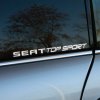 Na fotografii je biely nápis „SEAT Top Sport“ na bočnom okne auta. Písmo je elegantné, s jasnými, čistými líniami, ktoré pôsobia moderným dojmom a sú vhodné pre značenie športových modelov. Tento nápis je umiestnený na tmavom pozadí auta, čo umožňuje jeho jasné vyniknutie. V pozadí skla sa jemne odráža prirodzené prostredie s obrysmi stromov a oblohy, pridávajúc obrázku hĺbku a kontext. Tento jednoduchý, no štýlový dizajn zdôrazňuje spojenie automobilu SEAT s vysokým výkonom a športovým duchom.