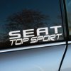 Na fotografii je zobrazený biely nápis „SEAT Top Sport“ na bočnom okne auta. Písmo je robustné a moderné, s ostrými hranami, čo zdôrazňuje športový charakter značky SEAT. Nápis je umiestnený na tmavom pozadí auta, vďaka čomu vyniká a zároveň pôsobí elegantne. V pozadí sa jemne odrážajú stromy a svetlomodrá obloha, čo dodáva obrázku prirodzený kontrast a zvýrazňuje čistotu a jednoduchosť dizajnu. Celkový dojem je súčasný a atraktívny, v súlade s imidžom značky SEAT ako výrobcu dynamických a výkonných automobilov.