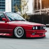 Červené BMW E30 M3 s upraveným nízkym podvozkom a ikonickými sieťovými diskami, zaparkované v modernom mestskom prostredí.