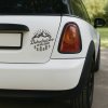Plotrovaná nálepka na auto s nápisom „Dobrodružstvo je môj život“, motív hôr, stromov a slnečných lúčov.