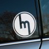 Plotrovaná nálepka s historickým logom značky Mazda, jednoduchý kruhový motív s písmenom M v minimalistickom spracovaní.