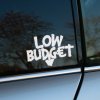 Plotrovaná nálepka s nápisom LOW BUDGET v originálnom ručne písanom štýle, doplnená malou šípkou smerujúcou nadol.