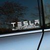 Plotrovaná nálepka s futuristickým písmom a akronymom TESLA – TOMORROW EXISTS SILENTLY LEADING AHEAD.