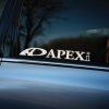 Plotrovaná nálepka na auto s logom APEXi, výrazný text doplnený ikonickým symbolom oblúka naľavo od nápisu.