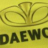 Detail potlače na reflexnej veste s logom Daewoo, plastický vejárový motív a hrubé čierne písmo na žltej tkanine.