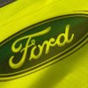 Detail potlače na reflexnej veste s logom Ford, oválny modrý motív s výrazným žltým písmom na reflexnej tkanine.