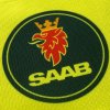 Detail potlače na veste s logom SAAB, červený lev s korunou v modrom kruhu a žltý nápis SAAB v spodnej časti.
