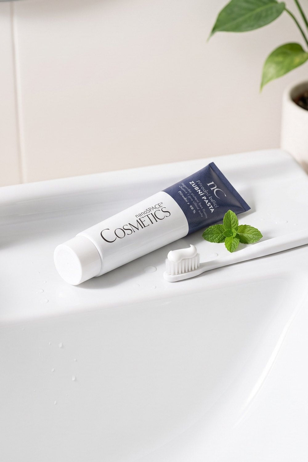 Tuba zubní pasty nanoSPACE Cosmetics a kartáček s pastou na okraji bílého umyvadla, lístek máty