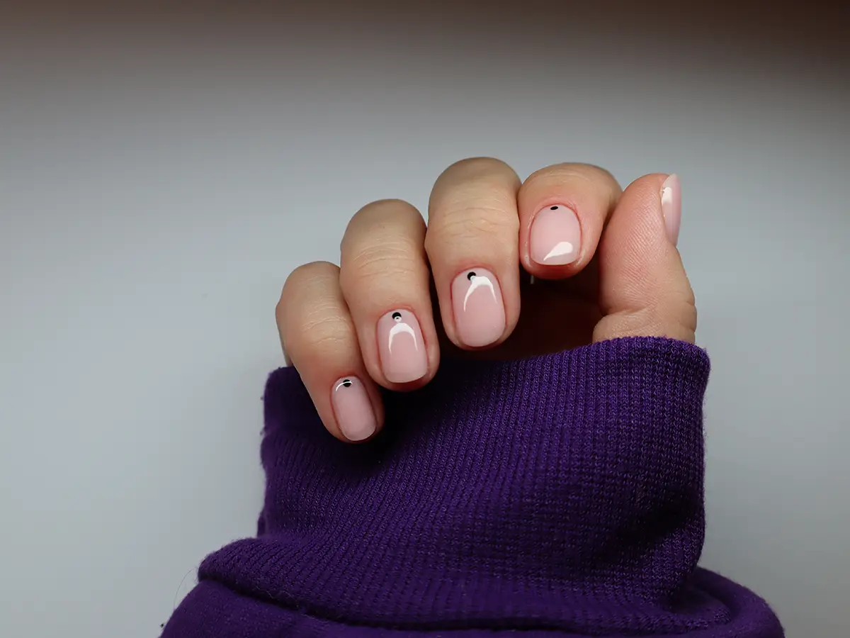 Manikúra Praha: ruka dívky ve fialovém svetru s krásnou nude manikúrou - růžovými nehty a černou tečkou.