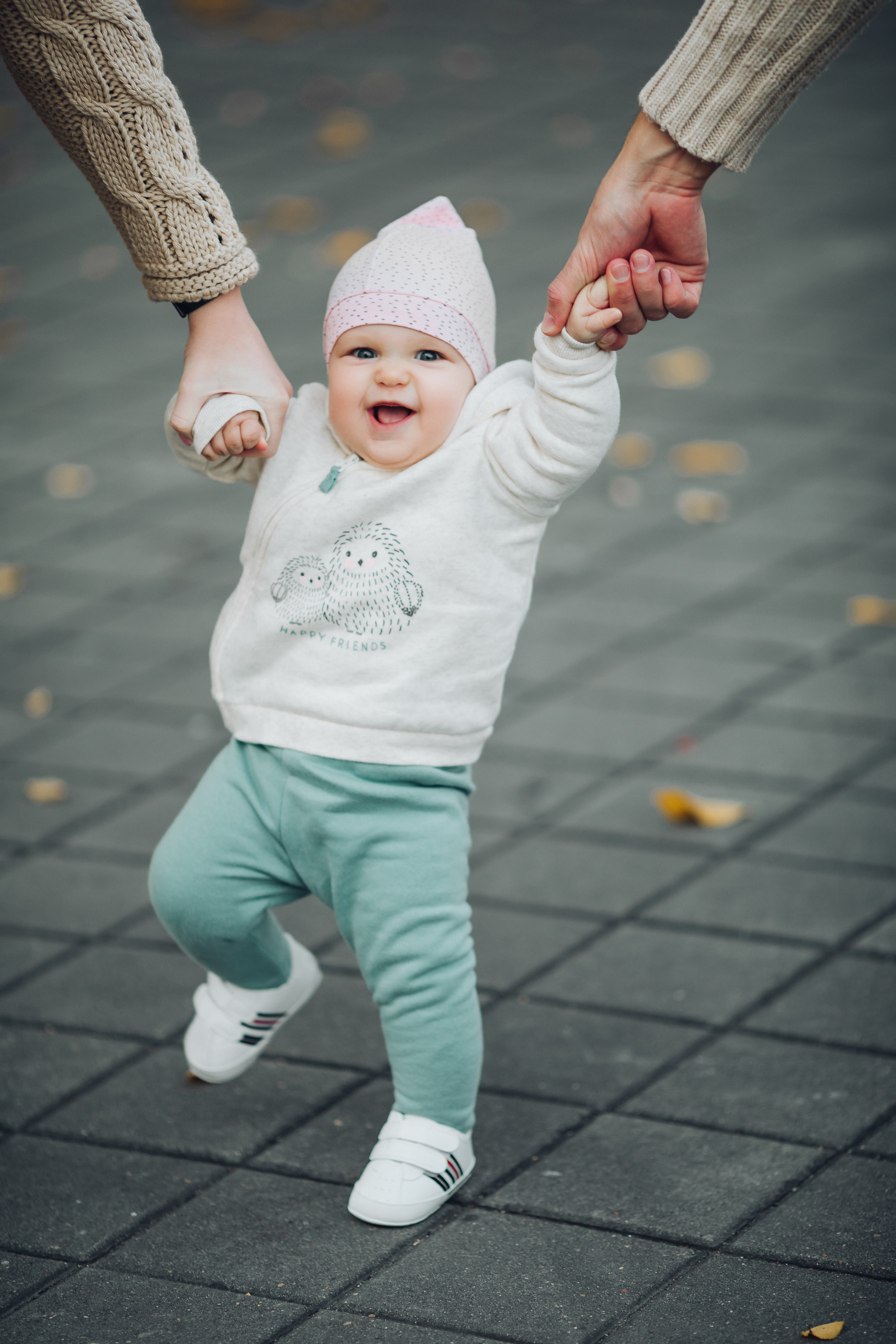 portrait-cute-sweet-baby-during-walking-outside-parents-holding-their-satisfied-child-by-hands-while-it-walking-happy-smiling-infant-white-sweater-green-pants-white-sneakers-doing-steps