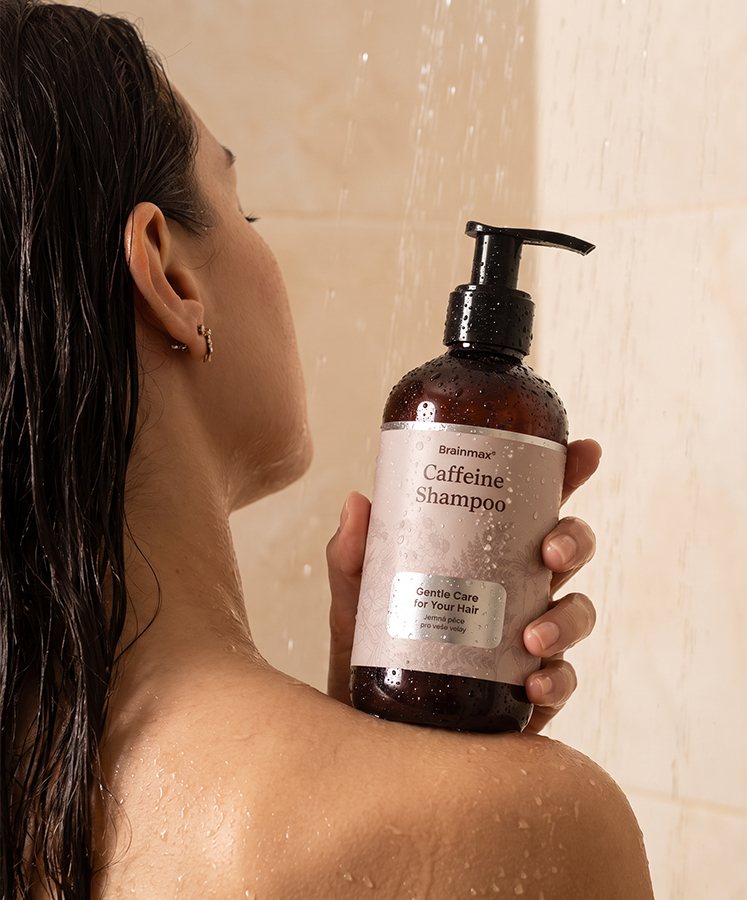 Žena ve sprše s lahví Brainmax Caffeine Shampoo na rameni