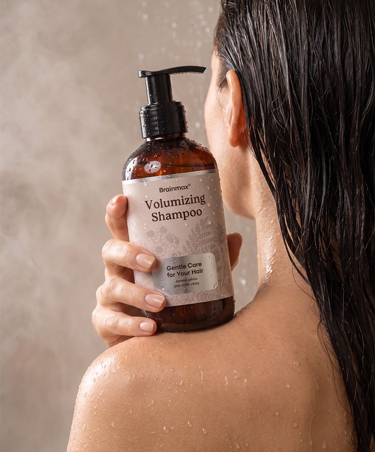 Žena ve sprše s lahví Brainmax Volumizing Shampoo na rameni