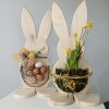 Dva maxi velikonoční zajíci s kapsou z pletiva. V jedné z nich je květina a mech, druhý má na krku žluto-modrou mašli a v košíku na bříšku přírodní, ale různobarevná vejce. Na bílém stole s šedým pozadím.