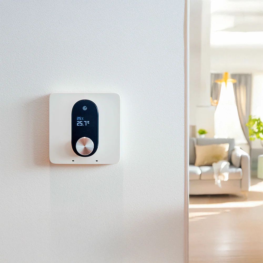 Bílý nástěnný termostat LinkedGo s kovovým otočným knoflíkem a displejem zobrazujícím vlhkost 25 % a teplotu 25,7 °C, v pozadí obývací pokoj