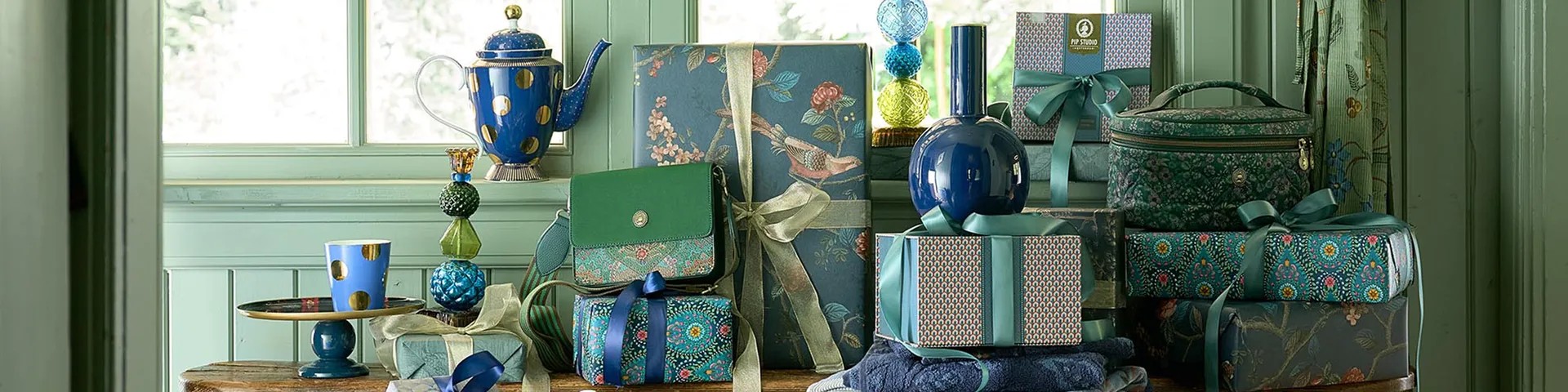 Tipy na luxusní dárky pro ženy značky Pip Studio. Na fotografii je designový modrý porcelán se zlatými detaily, elegantní zelená crossbody kabelka, kosmetický kufřík a dárkové krabice zabalené v luxusním balicím papíru s květinovými vzory a saténovými stuhami.