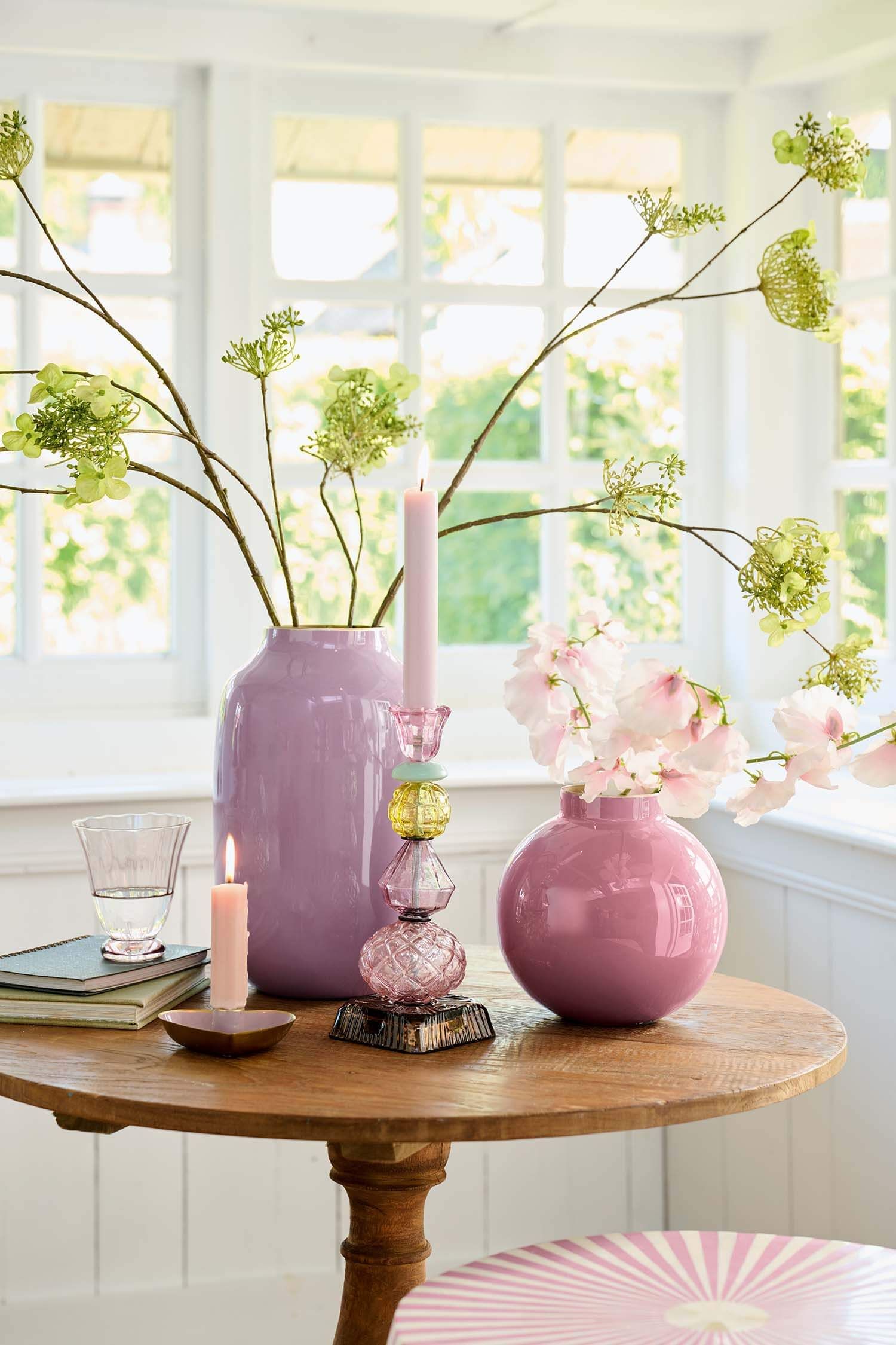 Velká lila fialová kovová váza Pip Studio Lily & Lotus a menší kulatá růžová váza s květinami a skleněným svícnem na dřevěném stolku v prosvětleném interiéru vedle proužkovaného kulatého pufu