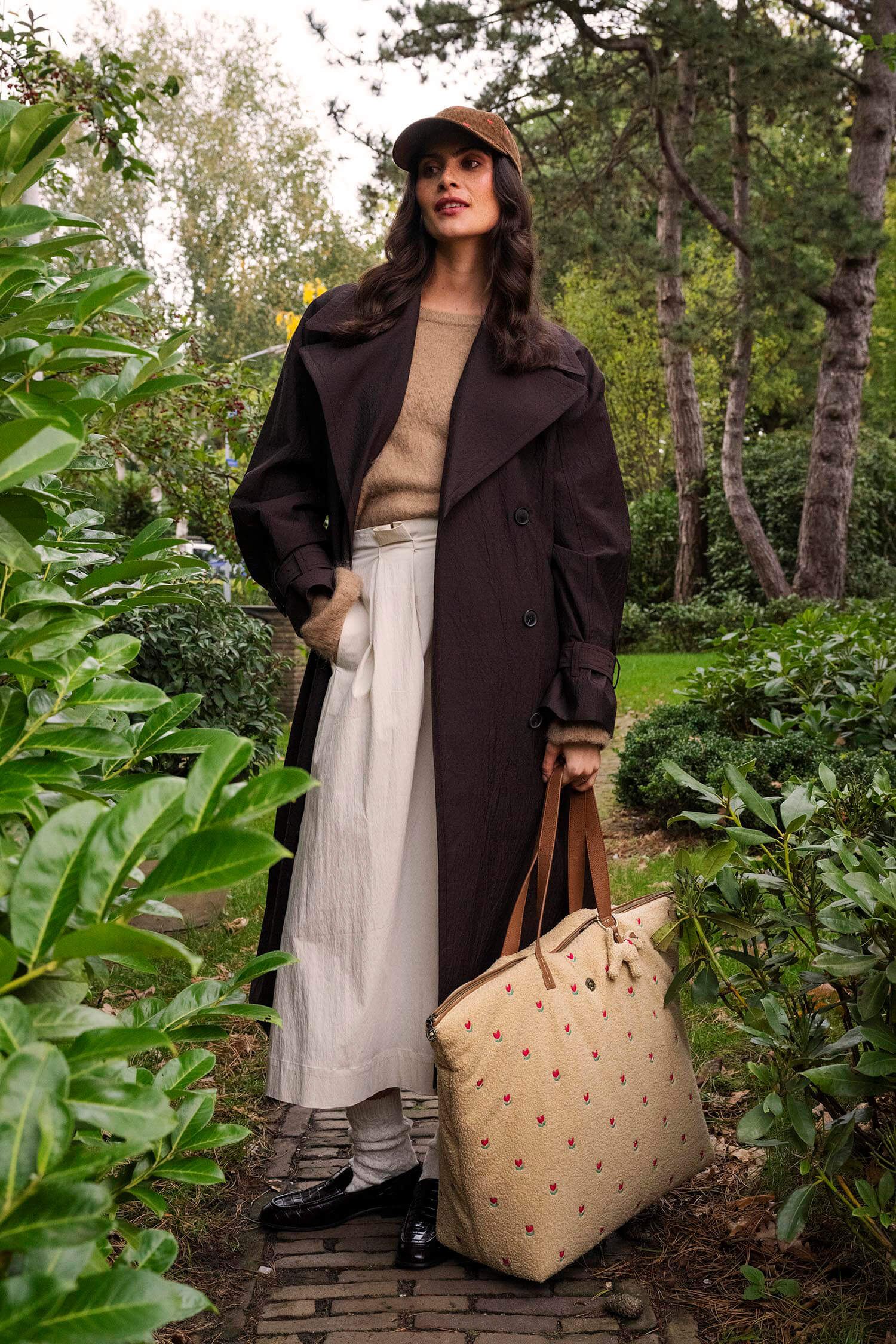 Pip Studio Tilda Tote Bag Teddy Sand velká cestovní taška, písková na modelce v tmavě hnědém kabátu a béžovém oblečení mezi rostlinami na kostkovaném chodniku