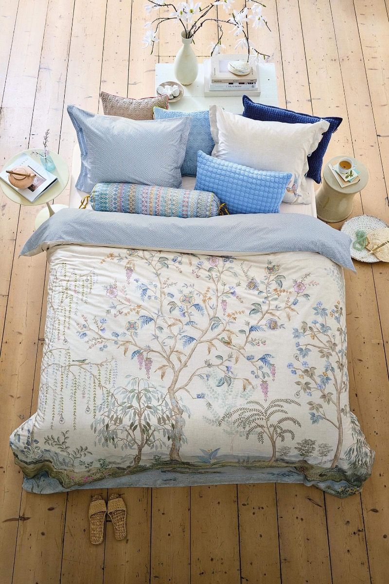 Produktová fotografie ložnice na dřevěné podlaze z e-shopu Pipa Ateliér představující povlečení Pip Studio Marakele Off-white s motivem vrb, exotických stromů a rajských ptáků. Postel zdobí pastelově modré dekorační a sametové polštáře. Inspirace pro barevné sladění ložnice, koupelny a kuchyně v České Budějovice.