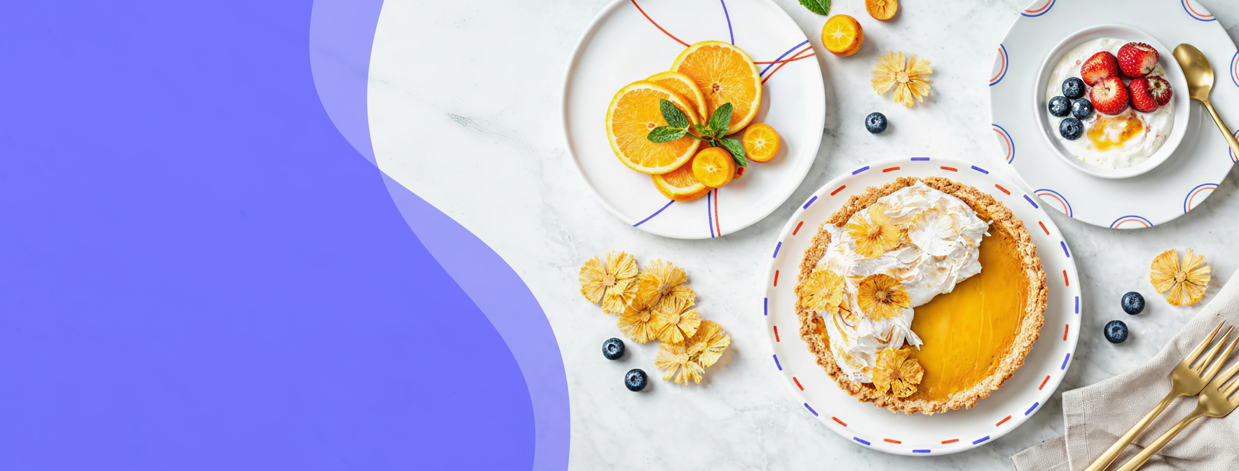 Kolekce Double Trouble – bílé talíře s modro-červeným dekorem, servírované s citrusovým koláčem, plátky pomeranče a jogurtem s ovocem na světlém podkladu s modrým grafickým přechodem.