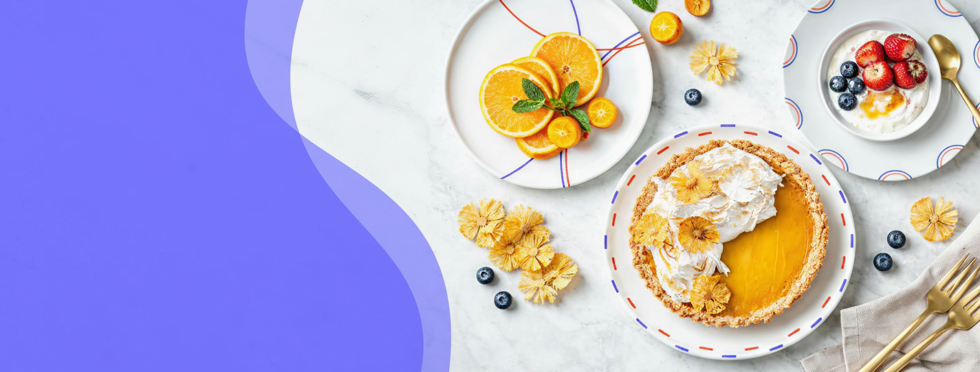 Kolekce Double Trouble – bílé talíře s modro-červeným dekorem, servírované s citrusovým koláčem, plátky pomeranče a jogurtem s ovocem na světlém podkladu s modrým grafickým přechodem.