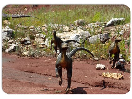 Dětské plastové prostírání s motivem malých dinosaurů v přírodní scenérii, na rudém podkladu s kamením a rostlinami, hravá dekorace na stůl.