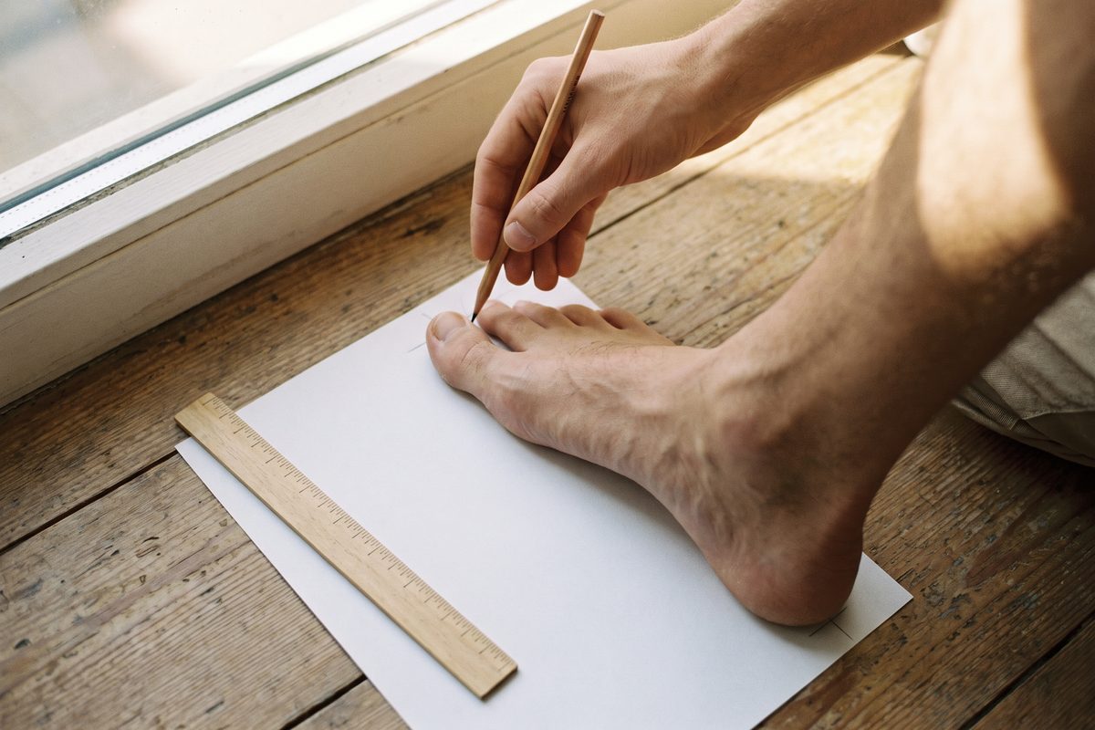 Muž si na papíře na podlaze obkresluje a měří délku chodidla