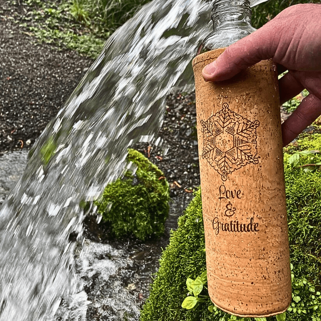 Hand holding a cork-sleeved ‘Love & Gratitude’ glass water bottle under a flowing stream in a mossy forest