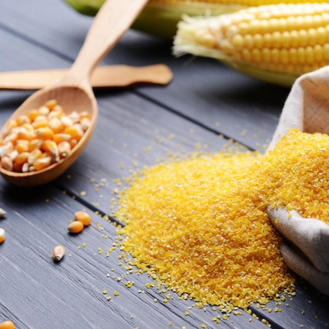 Corn grits and corn kernels on a wooden spoon, with a pile of yellow cornmeal on a dark table.