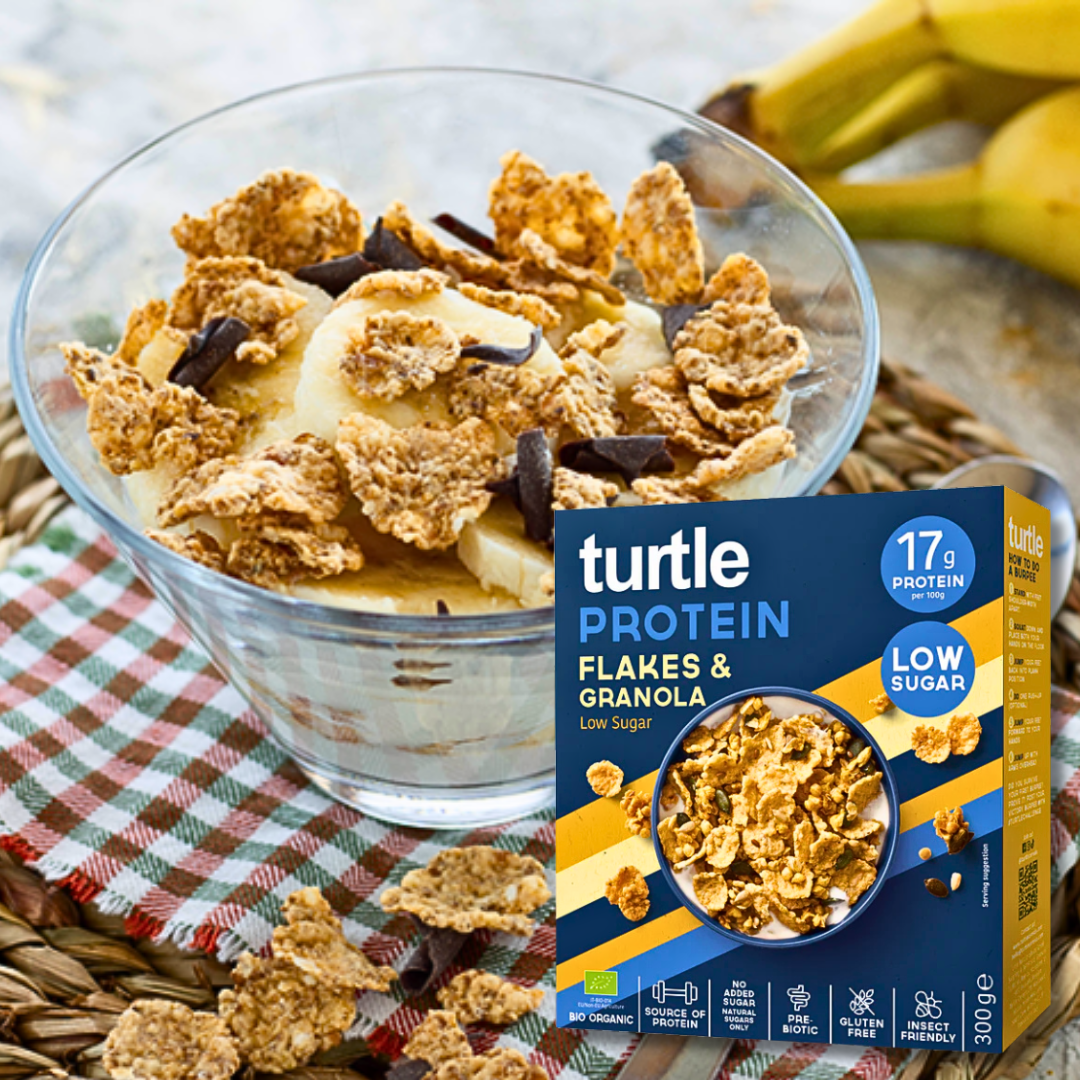 Glass bowl with Turtle Protein Flakes & Granola, banana slices and dark chocolate pieces with the cereal box in front.