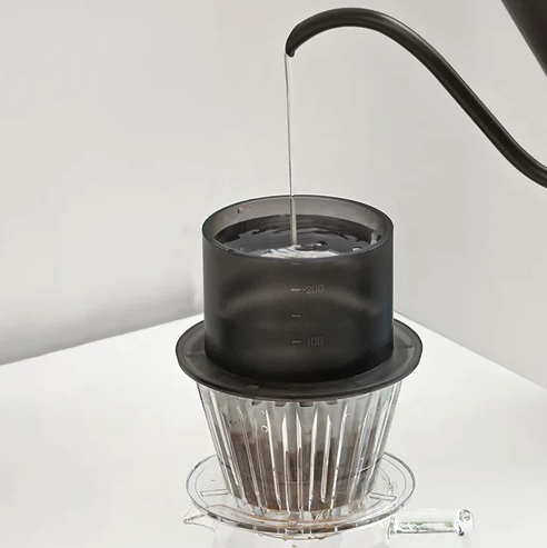 Nalévání horké vody z baristické konvice do sprchy umístěné na dripperu.