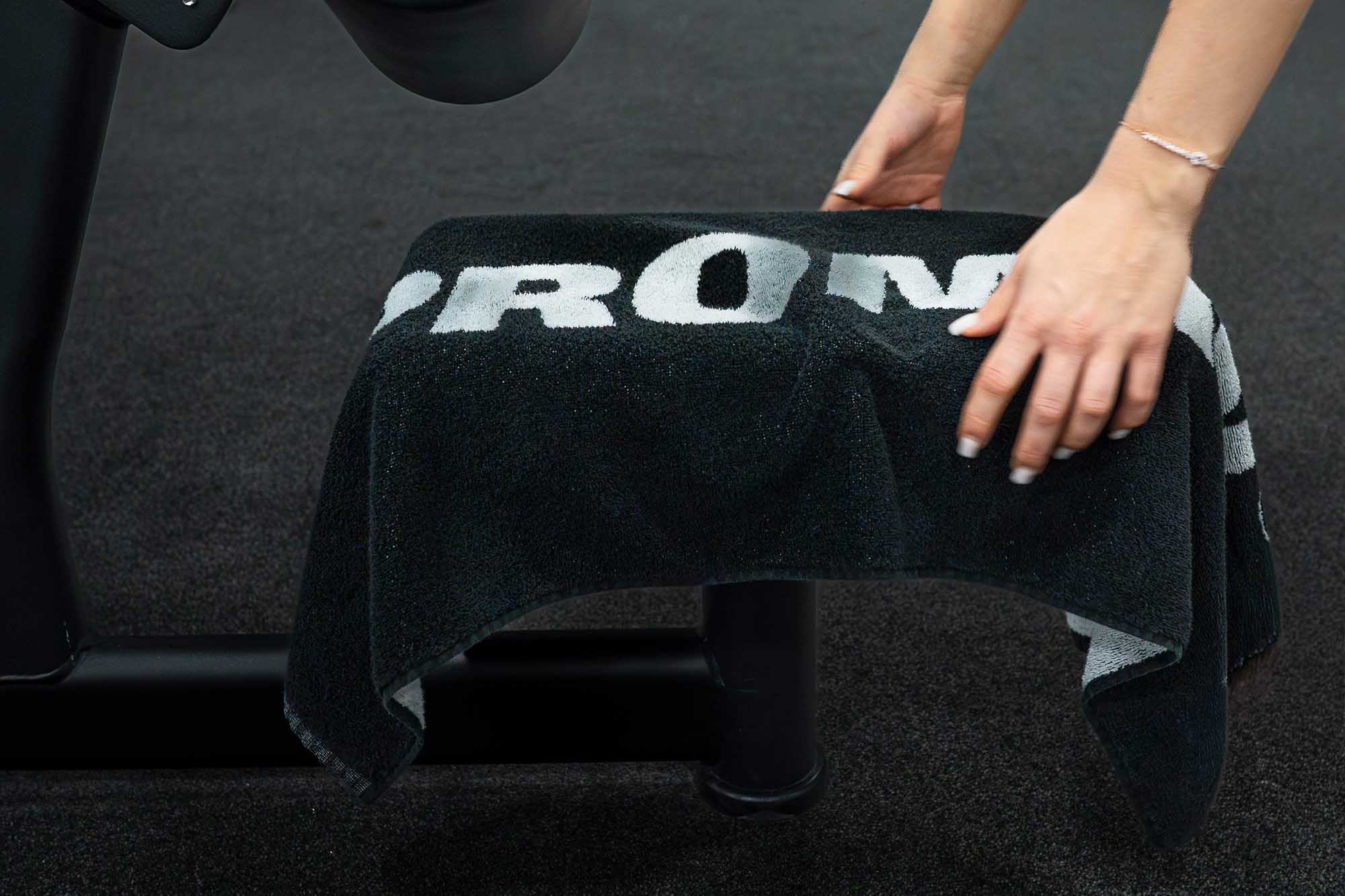 Černý fitness ručník PROM-IN položený na posilovacím stroji v posilovně.