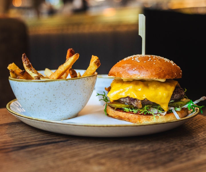 Burger s vyzrálým hovězím masem, čedarem, karamelizovanou cibulkou, nakládanou okurkou, salátem a zauzenými fermentovanými rajčaty, sezamová bulka, naše smažené hranolky
