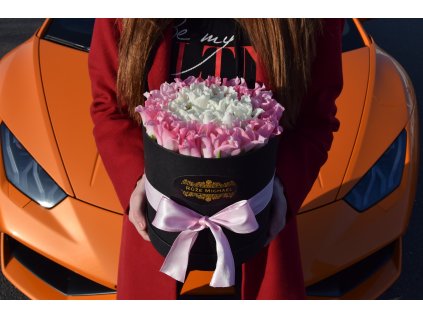 Elegantní černý box střední velikosti s jemně růžovými a bílými tulipány, před luxusním oranžovým sportovním vozem.