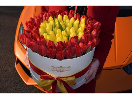 Luxusní box s tulipány v kombinaci rudých a žlutých květů, ozdobený zlatou a červenou stuhou, aranžovaný na kapotě oranžového sportovního auta.